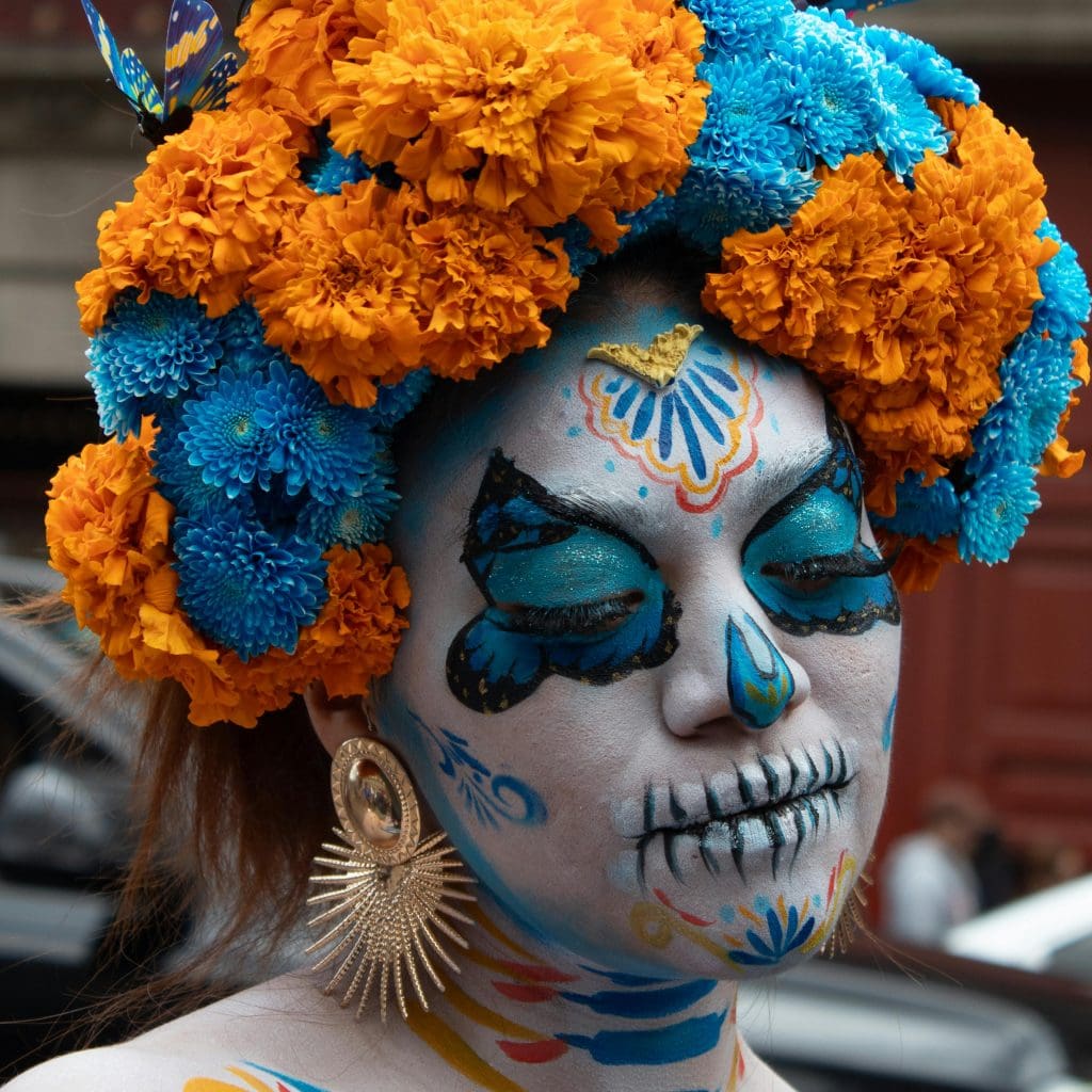 Image of woman wearing day of the dead skull makeup, blue and marigold flower crown, and large earrings